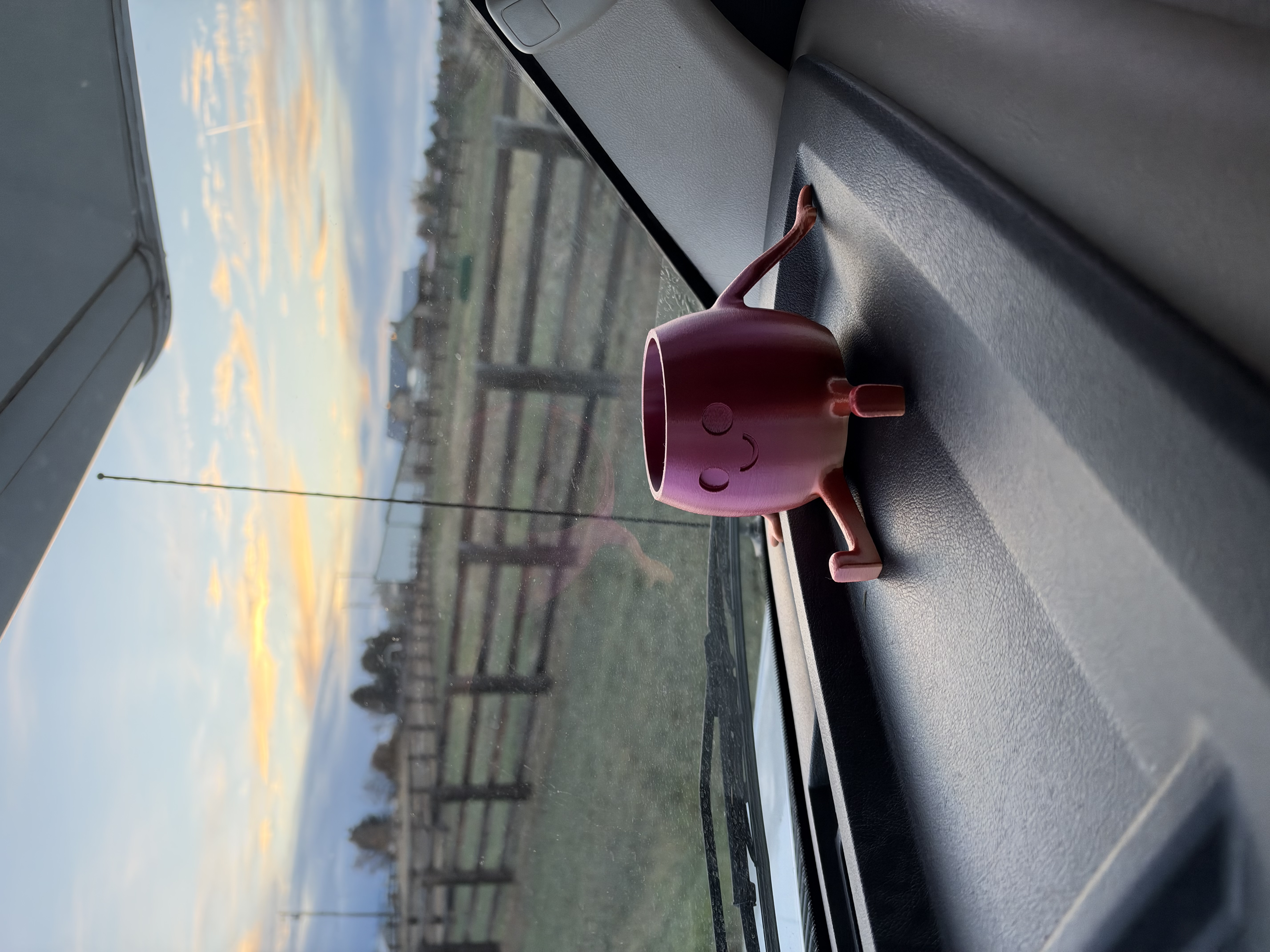 A smiling pink cup-shaped dashboard companion, with fences and sunset-colored clouds visible through the 23U windshield near Helena.